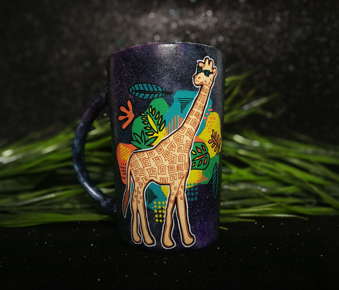 A Black galaxy hand painted mug with a giraffe standing wearing sunglasses and behind it colorful Hawaiian leaves in green, yellow and orange.