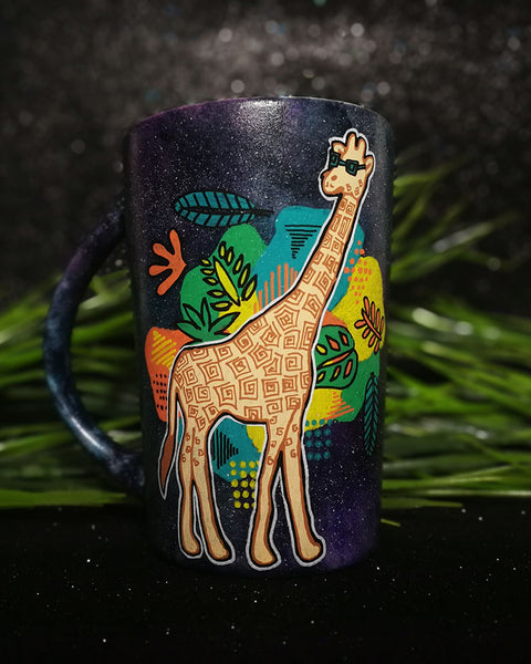 A Black galaxy hand painted mug with a giraffe standing wearing sunglasses and behind it colorful Hawaiian leaves in green, yellow and orange.