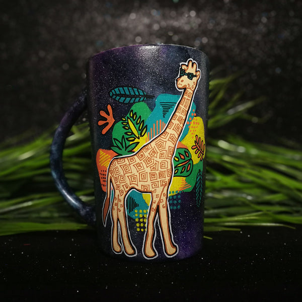 A Black galaxy hand painted mug with a giraffe standing wearing sunglasses and behind it colorful Hawaiian leaves in green, yellow and orange.