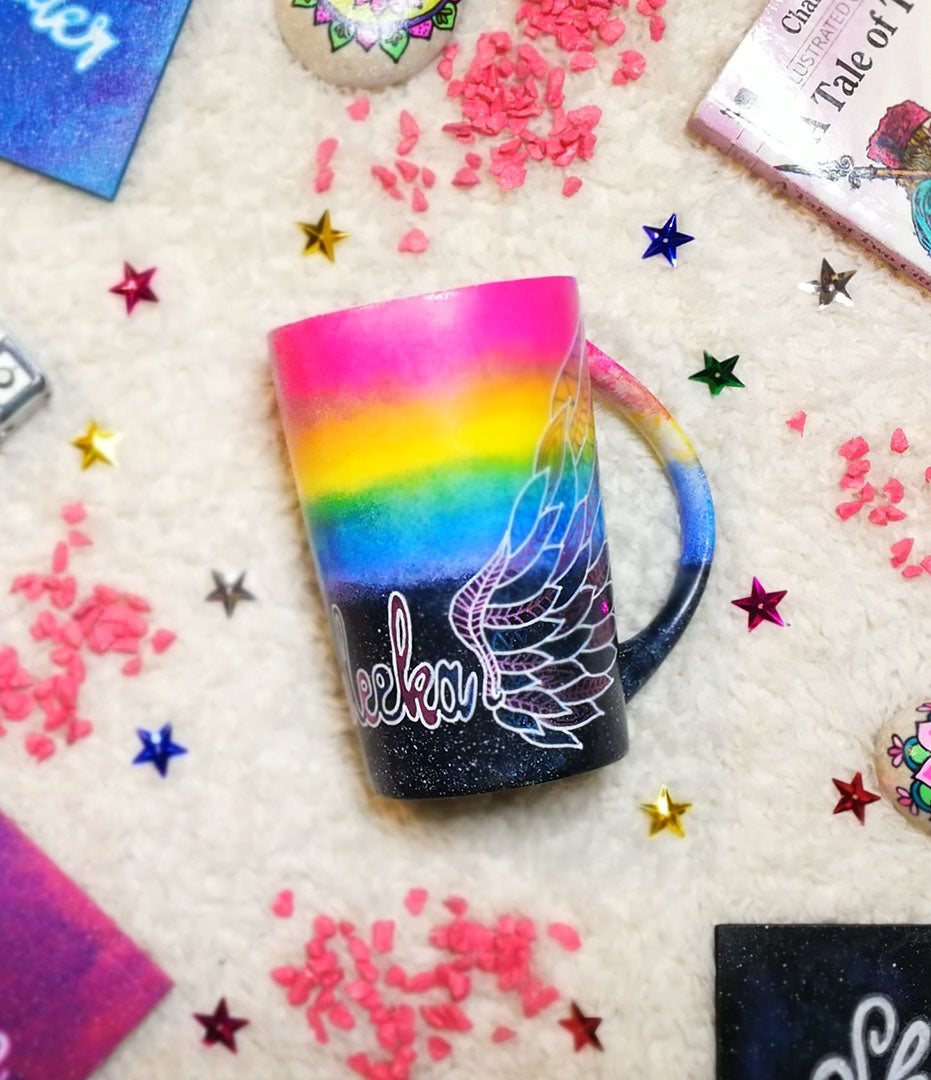 A gradient hand painted mug(rainbow colors in the top half and black galaxy in the bottom" with a wing drawn on one side 