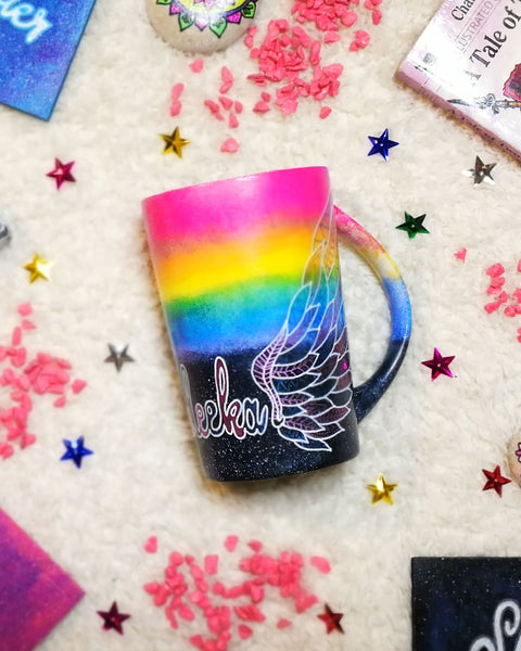 A gradient hand painted mug(rainbow colors in the top half and black galaxy in the bottom" with a wing drawn on one side 