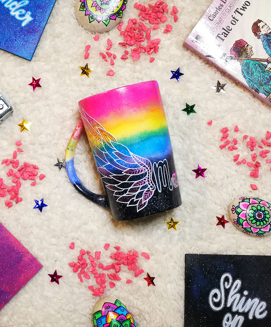 A gradient hand painted mug(rainbow colors in the top half and black galaxy in the bottom" with a wing drawn on one side 