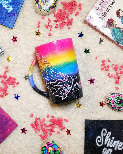 A gradient hand painted mug(rainbow colors in the top half and black galaxy in the bottom" with a wing drawn on one side 