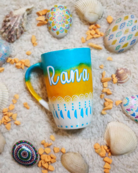 A gradient colored mug (from top blue then bej then white) with patterns on the bottom half horizontally in white and colored  with white and blue, and the name "rana" on the top