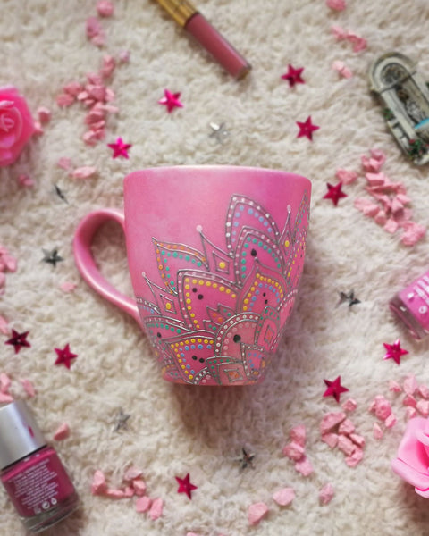 A Shimmery pink hand painted mug with a mandala drawn in the bottom center spread to both sides using a silver relief and dot art of mint, yellow, baby blue, pastel orange, grey, white and baby pink