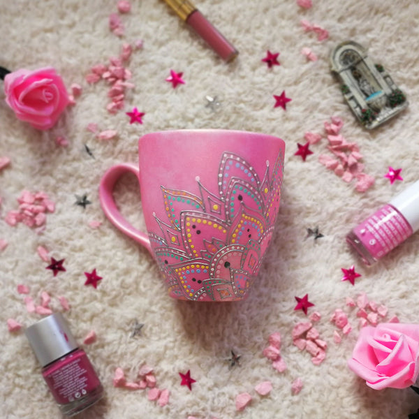 A Shimmery pink hand painted mug with a mandala drawn in the bottom center spread to both sides using a silver relief and dot art of mint, yellow, baby blue, pastel orange, grey, white and baby pink