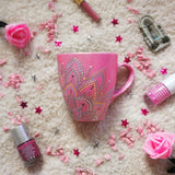 A Shimmery pink hand painted mug with a mandala drawn in the bottom center spread to both sides using a silver relief and dot art of mint, yellow, baby blue, pastel orange, grey, white and baby pink