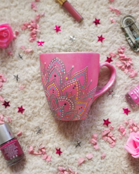 A Shimmery pink hand painted mug with a mandala drawn in the bottom center spread to both sides using a silver relief and dot art of mint, yellow, baby blue, pastel orange, grey, white and baby pink