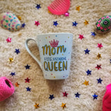 A baby Blue mug with the word MOM with a pink crown on top and Queen written in large font in gold and in between the words 