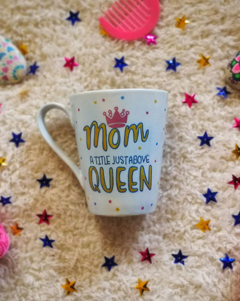 A baby Blue mug with the word MOM with a pink crown on top and Queen written in large font in gold and in between the words " a title just above". with gold , pink and blue dots scattered all over