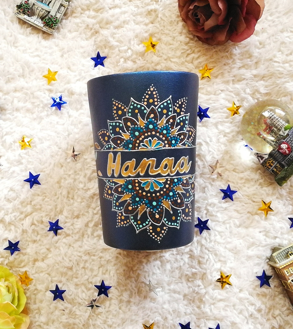 A Shimmery blue hand painted mug with a mandala split into 2 with a space in between that has the name "Hanaa" and the mandala is outlined with silver relief and dot art of gold, turquoise, bronze and bej