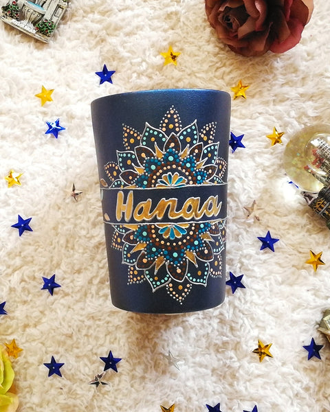 A Shimmery blue hand painted mug with a mandala split into 2 with a space in between that has the name "Hanaa" and the mandala is outlined with silver relief and dot art of gold, turquoise, bronze and bej