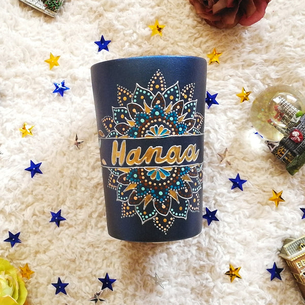 A Shimmery blue hand painted mug with a mandala split into 2 with a space in between that has the name "Hanaa" and the mandala is outlined with silver relief and dot art of gold, turquoise, bronze and bej