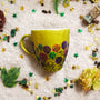 A Bronze hand painted mug with flowers drawn at the bottom half all around with black outliner and colored in purple, green,yellow and dark pink