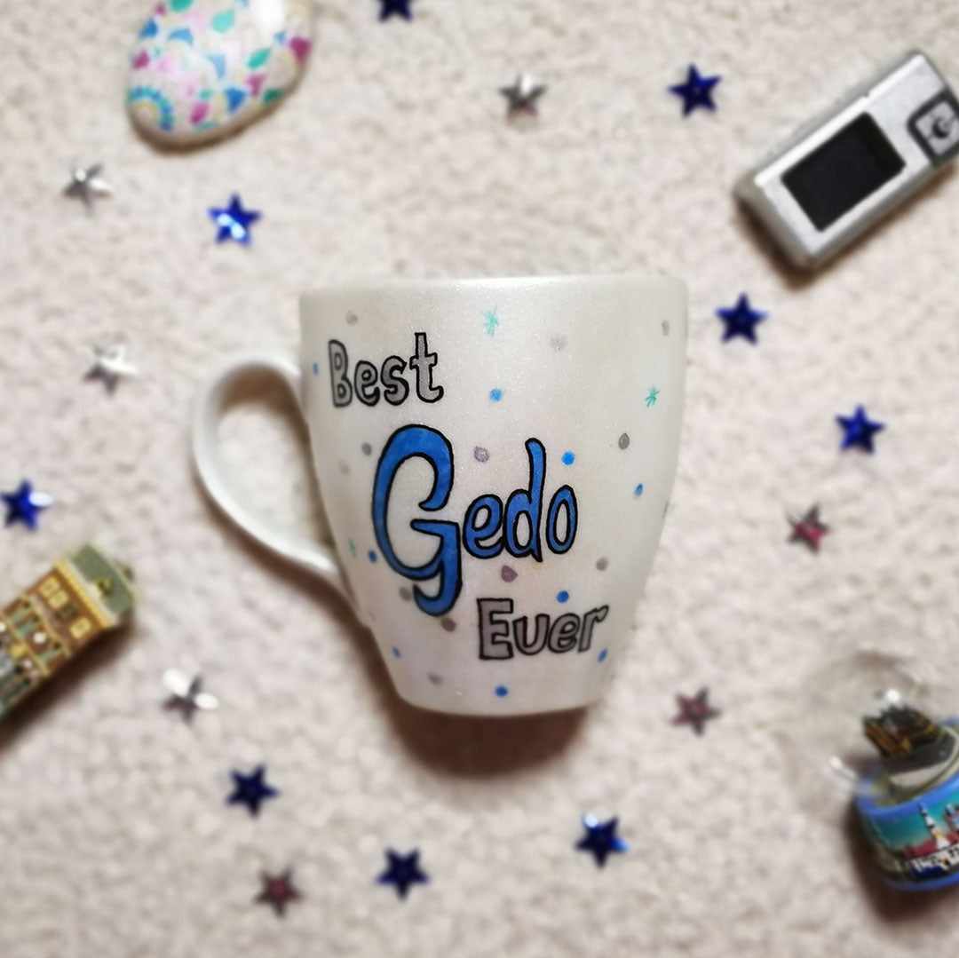 A hand painted pearl white mug with the words "best Gedo ever" and the word Gedo is in blue while the other in silver, with silver blue dots and green stars scattered all over