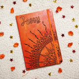 An orange yellow hand painted notebook with a quarter mandala drawn from the corner and the name 