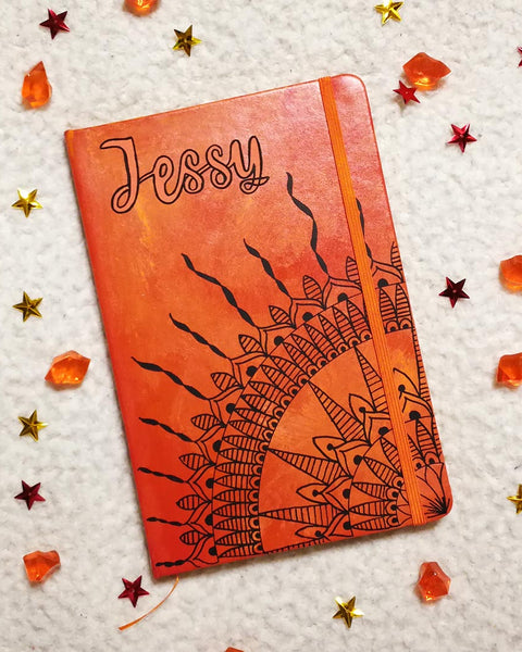 An orange yellow hand painted notebook with a quarter mandala drawn from the corner and the name "Jessy" written on top