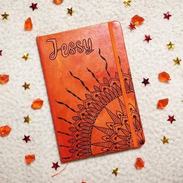 An orange yellow hand painted notebook with a quarter mandala drawn from the corner and the name "Jessy" written on top