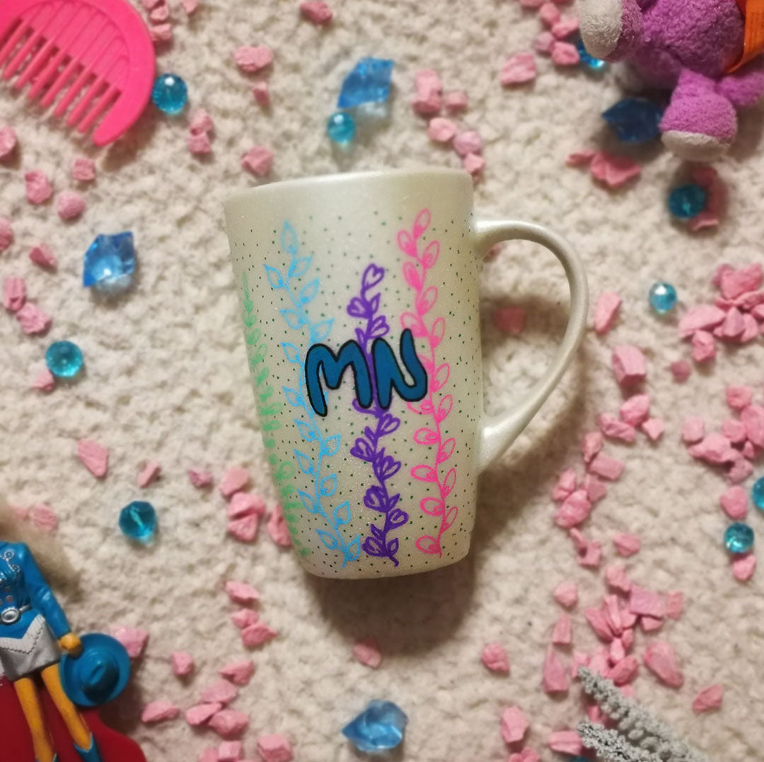 A pearl white hand painted mug with leaves draw on it vertically all over the mug from bottom to the top on colors of pink, green, blue, purple, orange and yellow with the letters "MN" written in the center