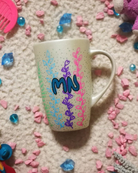 A pearl white hand painted mug with leaves draw on it vertically all over the mug from bottom to the top on colors of pink, green, blue, purple, orange and yellow with the letters "MN" written in the center