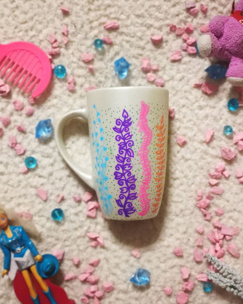 A pearl white hand painted mug with leaves draw on it vertically all over the mug from bottom to the top on colors of pink, green, blue, purple, orange and yellow