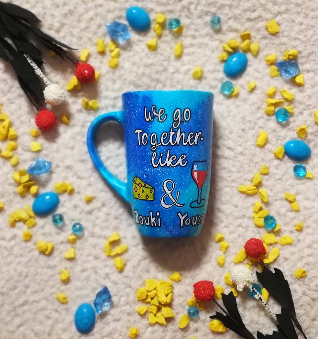 A blue galaxy hand painted mug with the sentence " we go together like" and then a triangular cheese and a cup of wine are drawn with the word "and" between them and under the cheese the name " Zouki" is written and under the wine the name " Youssy" is written