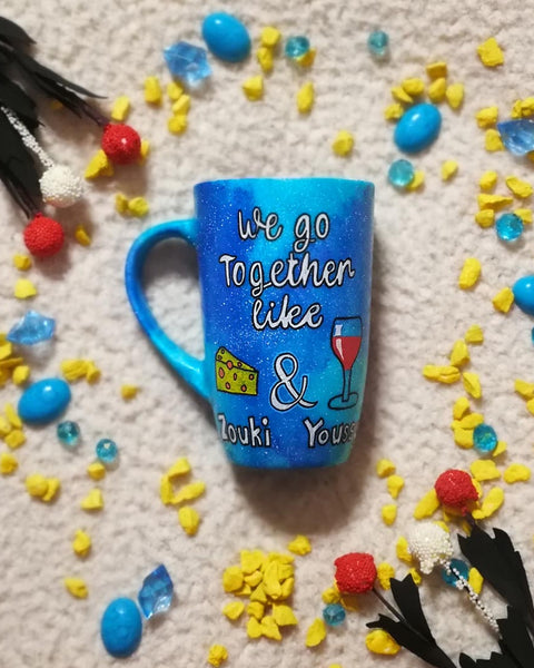 A blue galaxy hand painted mug with the sentence " we go together like" and then a triangular cheese and a cup of wine are drawn with the word "and" between them and under the cheese the name " Zouki" is written and under the wine the name " Youssy" is written