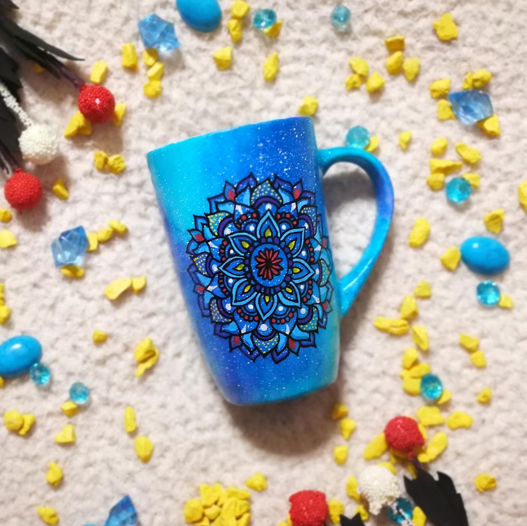 A hand painted blue galaxy mug with a mandala drawn on it in colors of red yellow and blue