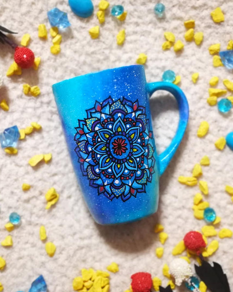 A hand painted blue galaxy mug with a mandala drawn on it in colors of red yellow and blue