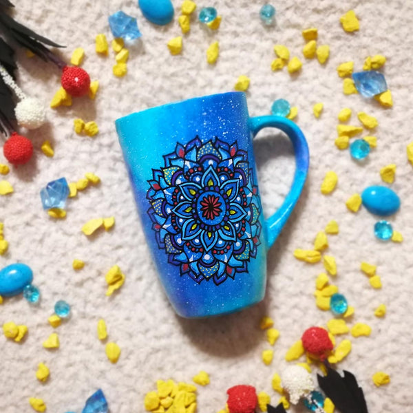 A hand painted blue galaxy mug with a mandala drawn on it in colors of red yellow and blue