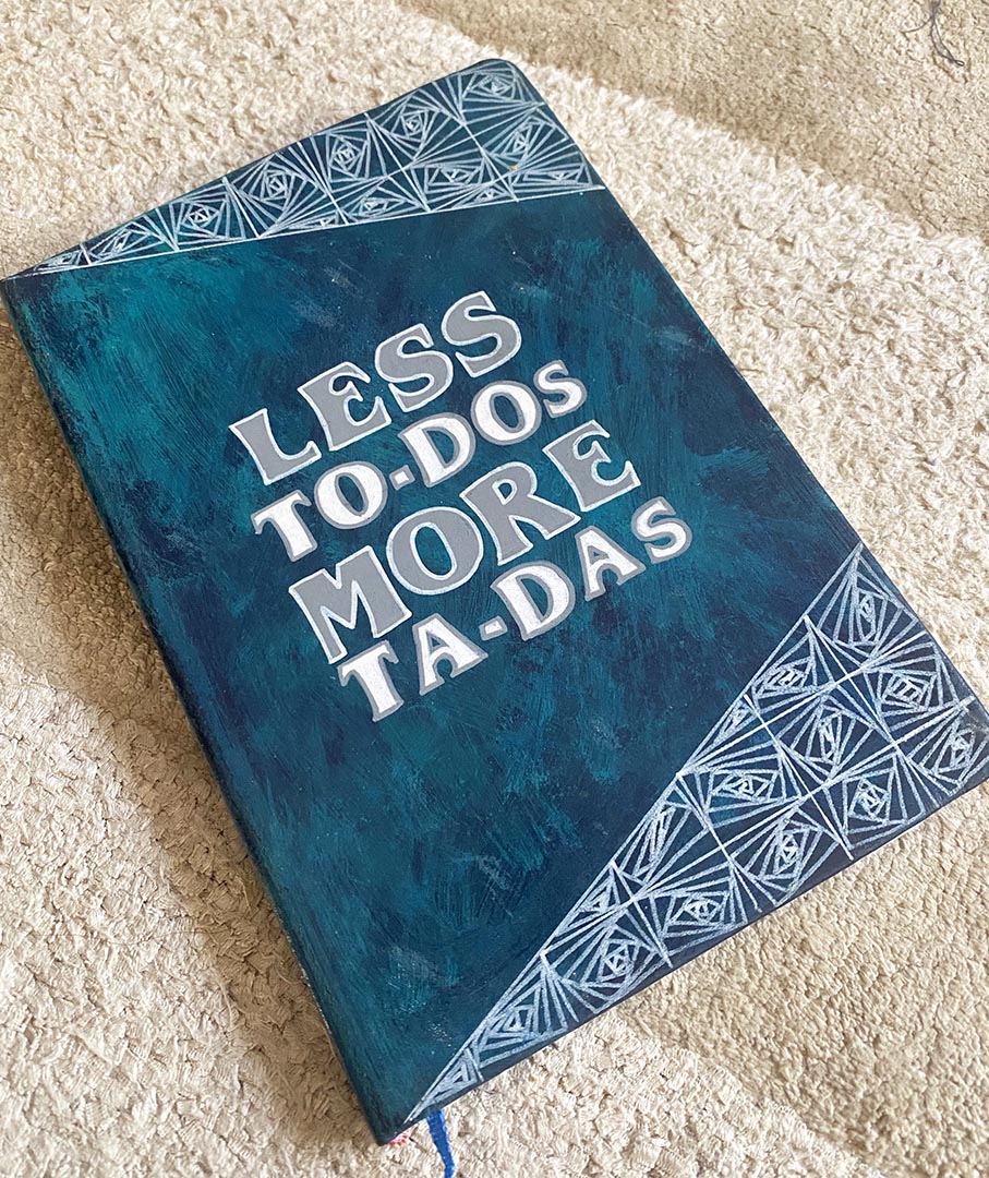 A Teal hand painted notebook witht he words "less to-dos more ta-das" written in the center and patterns on the top and bottom