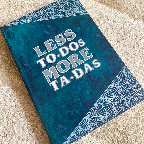 A Teal hand painted notebook witht he words "less to-dos more ta-das" written in the center and patterns on the top and bottom