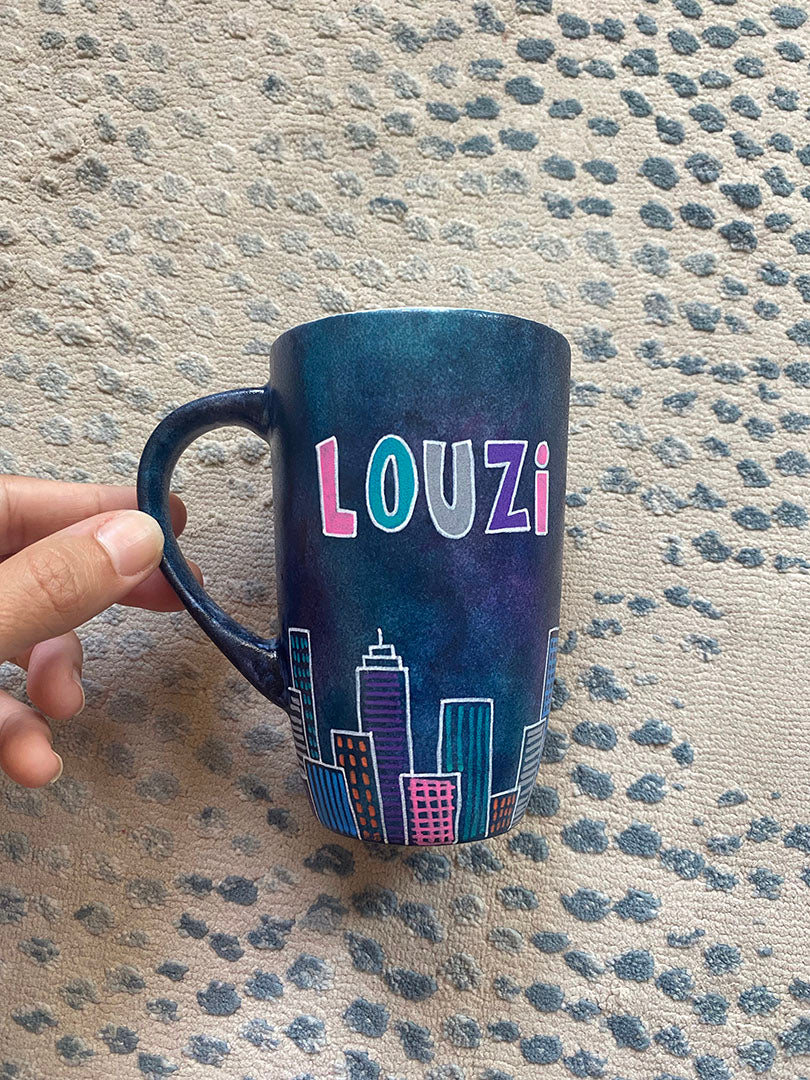 A Dark Sky handpainted mug with buildings drawn all over at the bottom half and the name "Louzi""