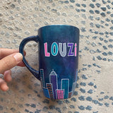 A Dark Sky handpainted mug with buildings drawn all over at the bottom half and the name 