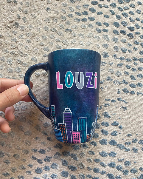 A Dark Sky handpainted mug with buildings drawn all over at the bottom half and the name "Louzi""