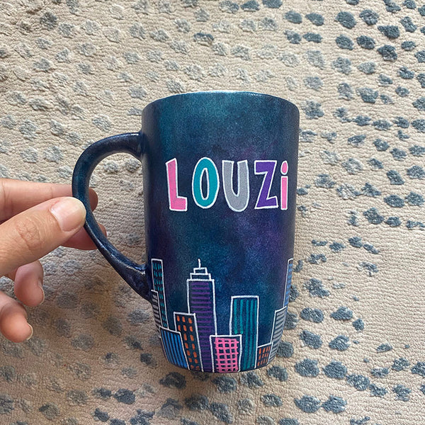 A Dark Sky handpainted mug with buildings drawn all over at the bottom half and the name "Louzi""