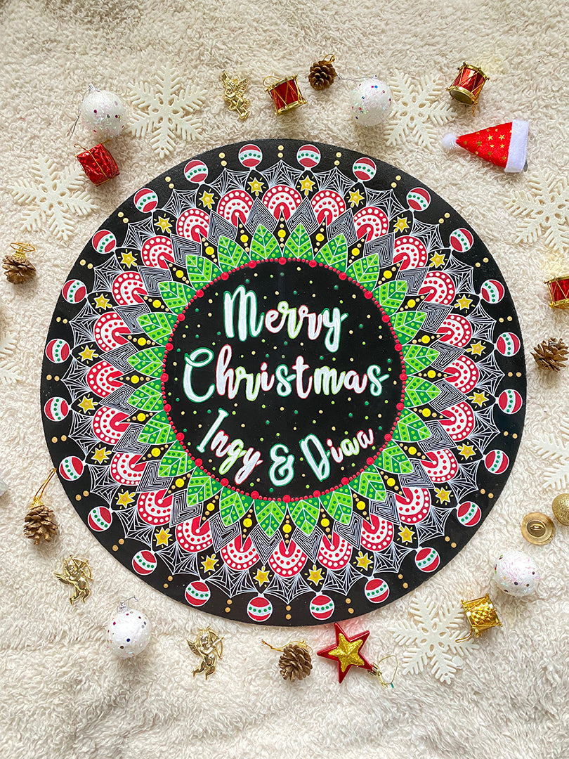 A Large 50cm in diameter mdf wooden plate, painted in black with a mandala drawn in white colored in green red and gold. and in the center the words "Merry christmas " written above and the names "Ingy and Diaa" written below