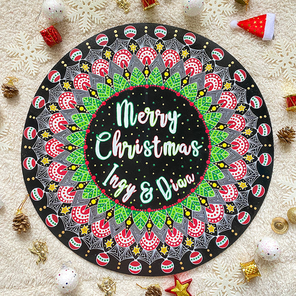 A Large 50cm in diameter mdf wooden plate, painted in black with a mandala drawn in white colored in green red and gold. and in the center the words "Merry christmas " written above and the names "Ingy and Diaa" written below