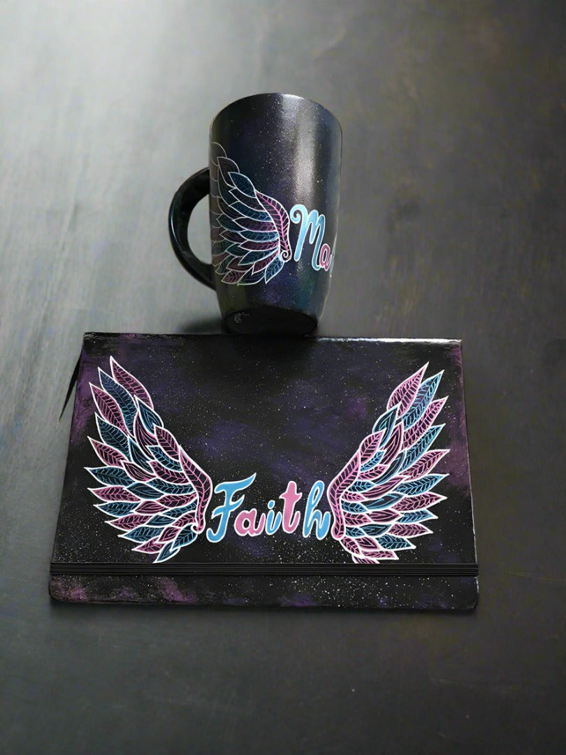 A black galaxy hand painted mug and notebook with a wings drawn on it and in between the mug has the name malak and the notebook has the word Faith