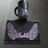 A black galaxy hand painted mug and notebook with a wings drawn on it and in between the mug has the name malak and the notebook has the word Faith