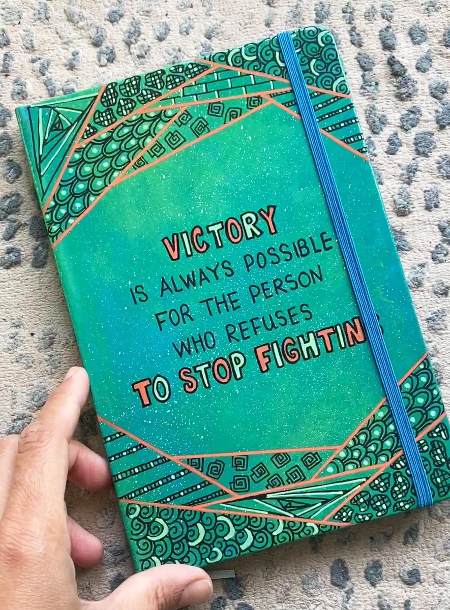 A green Galaxy notebook with patterns above and below and the words "Victory, is always for the person who refuses to stop fighting"