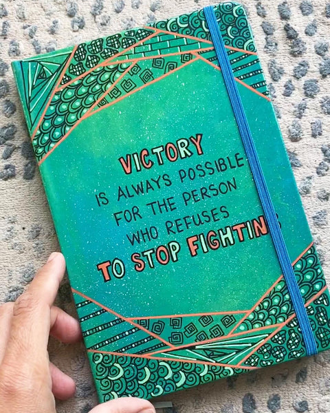 A green Galaxy notebook with patterns above and below and the words "Victory, is always for the person who refuses to stop fighting"