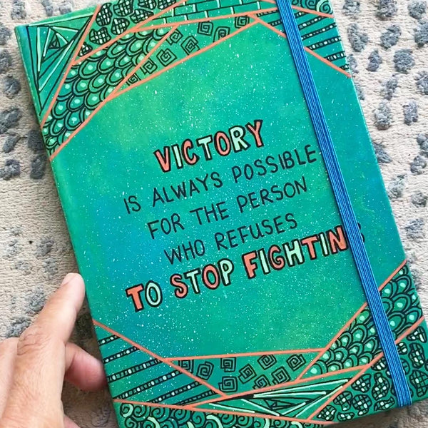 A green Galaxy notebook with patterns above and below and the words "Victory, is always for the person who refuses to stop fighting"