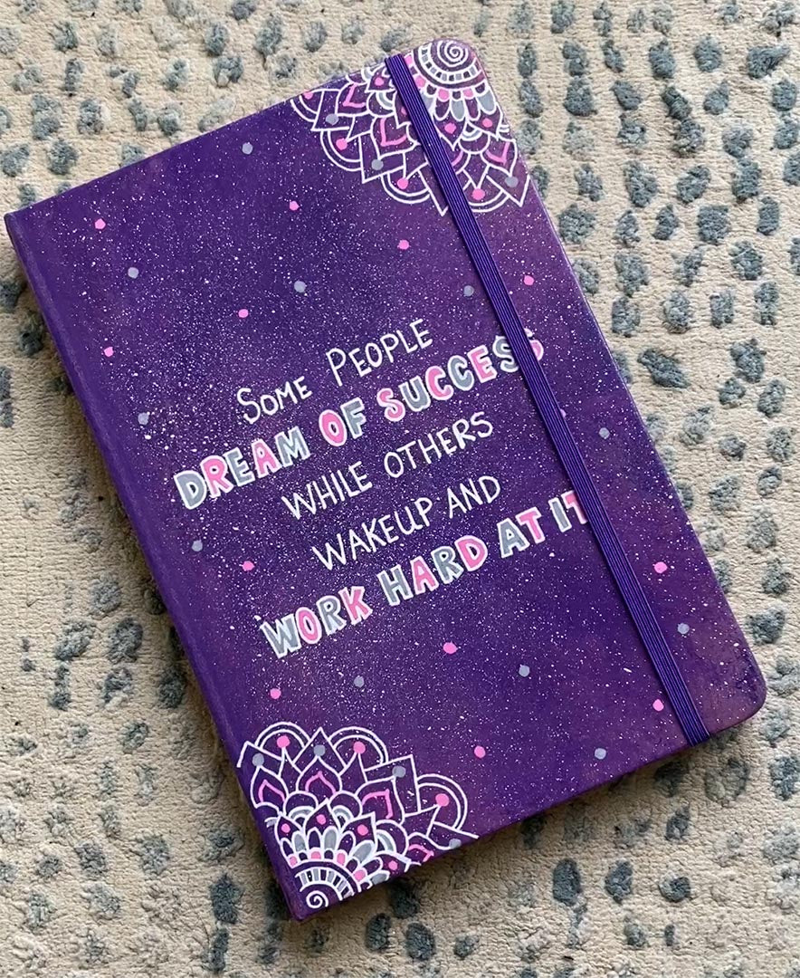 A Purple Galaxy Hand painted notebook with 2 small mandala drawn in white in the opposite corners and the quote written in the center "Some people Dream of success, while others wakeup and work hard at it"