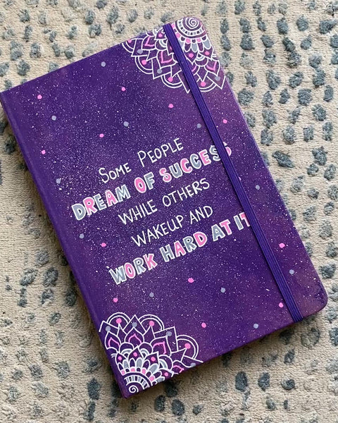 A Purple Galaxy Hand painted notebook with 2 small mandala drawn in white in the opposite corners and the quote written in the center "Some people Dream of success, while others wakeup and work hard at it"