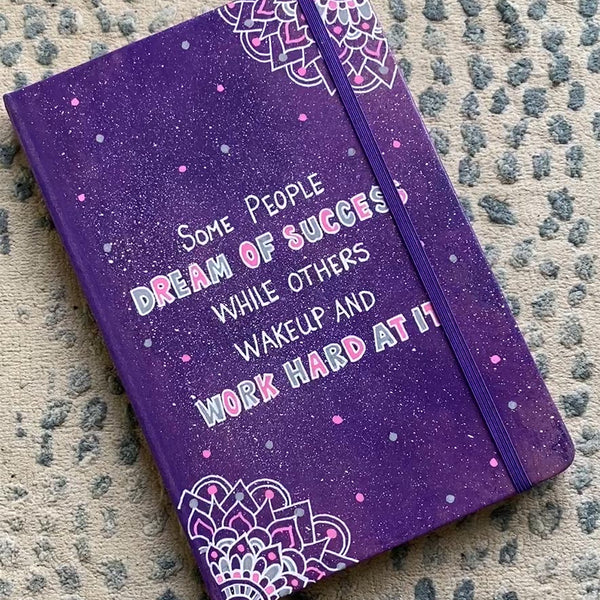 A Purple Galaxy Hand painted notebook with 2 small mandala drawn in white in the opposite corners and the quote written in the center "Some people Dream of success, while others wakeup and work hard at it"