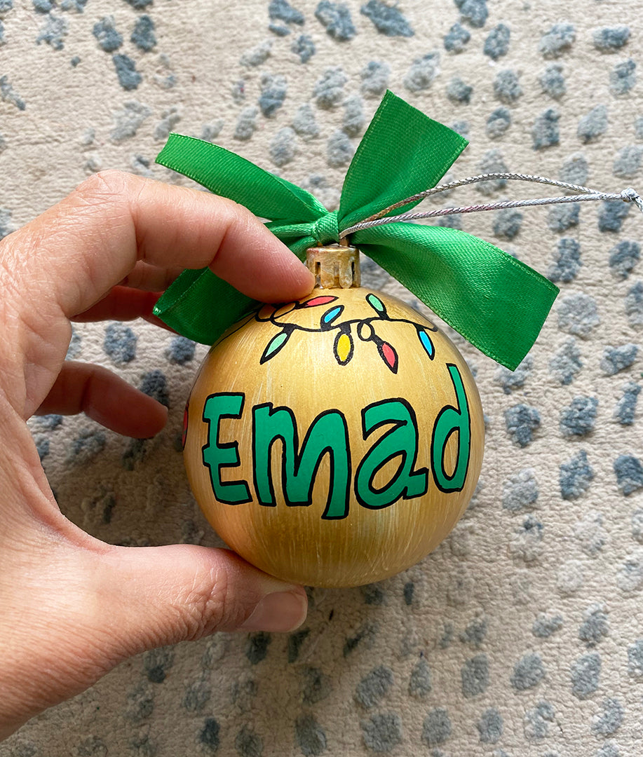 A gold hand painted ball ornament with festive lights all around the top and the name "Emad" written in green outlined with black