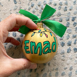 A gold hand painted ball ornament with festive lights all around the top and the name 