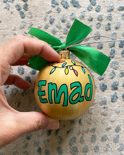 A gold hand painted ball ornament with festive lights all around the top and the name "Emad" written in green outlined with black