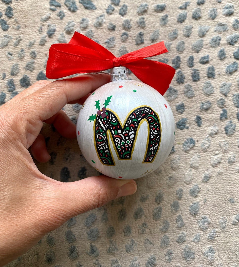 A Pearl white hand painted ball ornament with the letter "M" drawn filled with black doodles and colored with green and red, outlined with gold and green  and red dots scattered all over the ball with gold stars